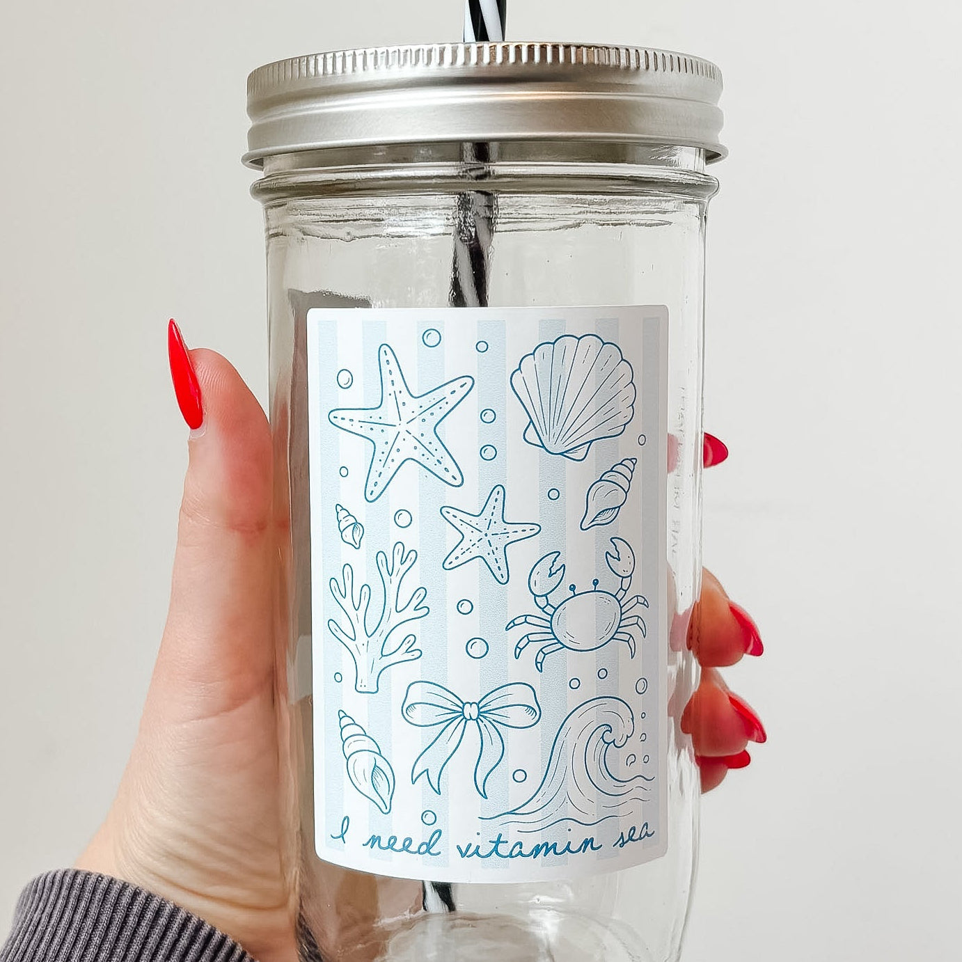 Hand holding mason jar tumbler that reads i need vitamin sea with starfish, shell, crab, coral and other ocean designs on pale blue and white striped background