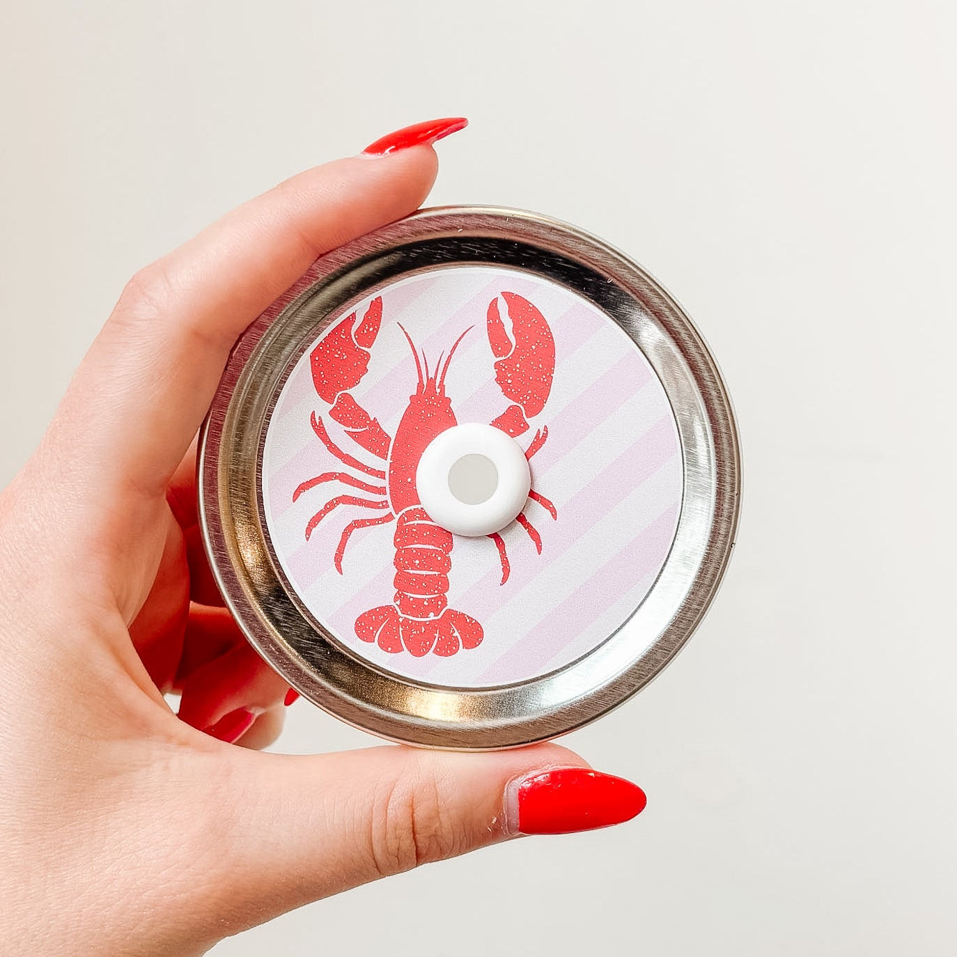 Hand holding mason jar patterned straw lid up close that has red lobster design on pale pink and white diagonal stripe background