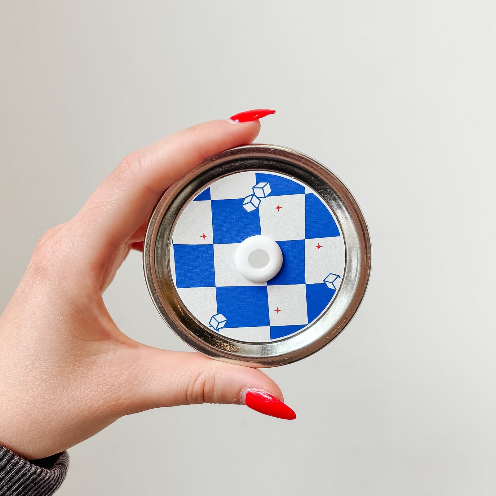 Hand holding a mason jar patterned straw lid that has blue and white checkered design with little iced cubes and tiny red stars