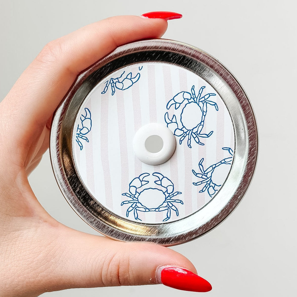 Hand holding mason jar patterned straw lid up close that has blue crab designs on pale pink and white striped background