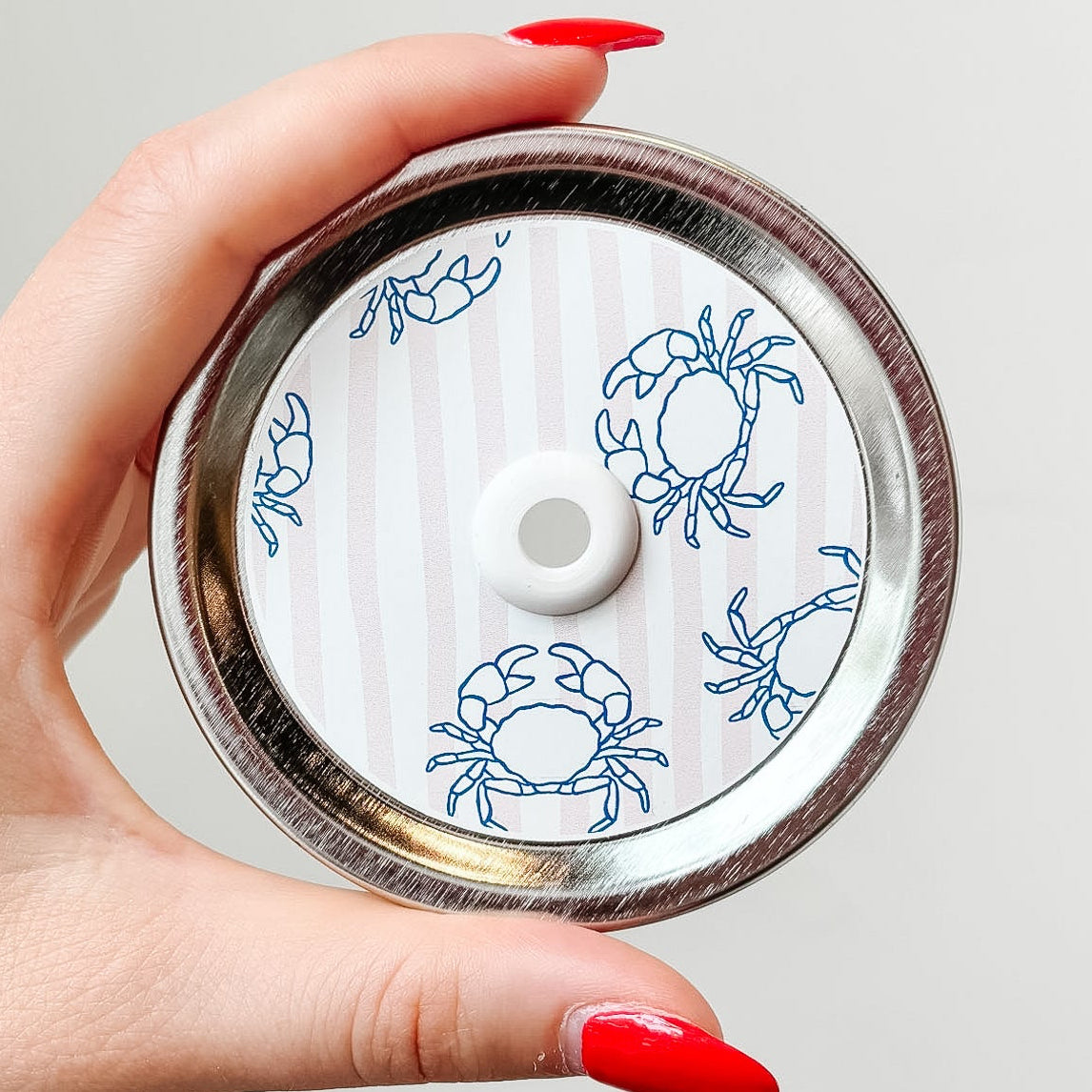 Hand holding mason jar patterned straw lid up close that has blue crab designs on pale pink and white striped background