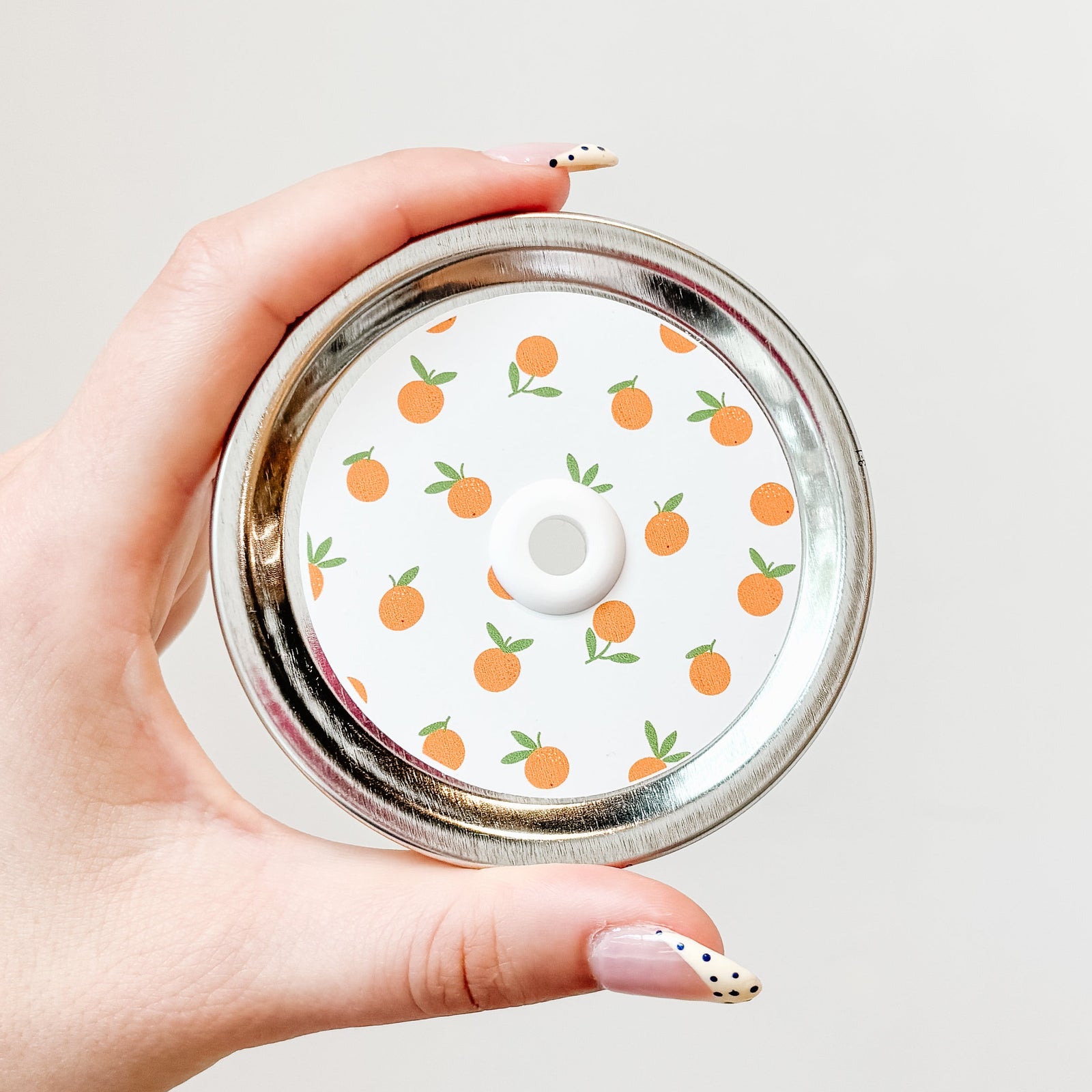 Hand holding a mason jar patterned straw lid with little oranges on a white background.