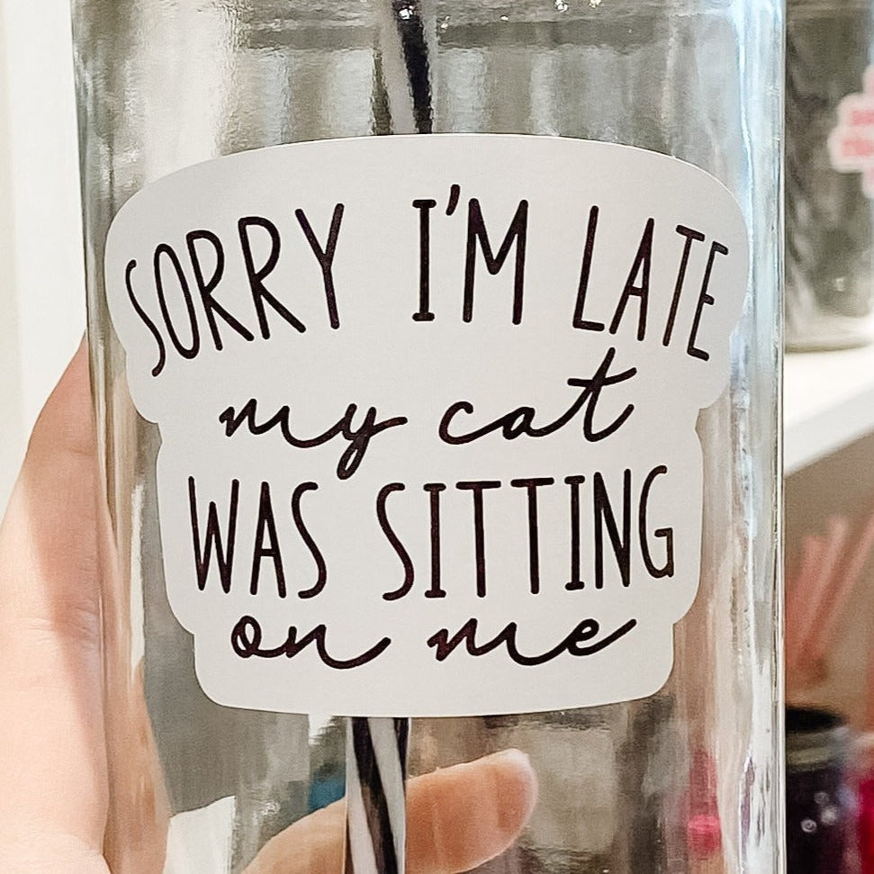 Close up of a hand holding a mason jar tumbler that says “Sorry I’m late my cat was sitting on me” in black letters on a white background.