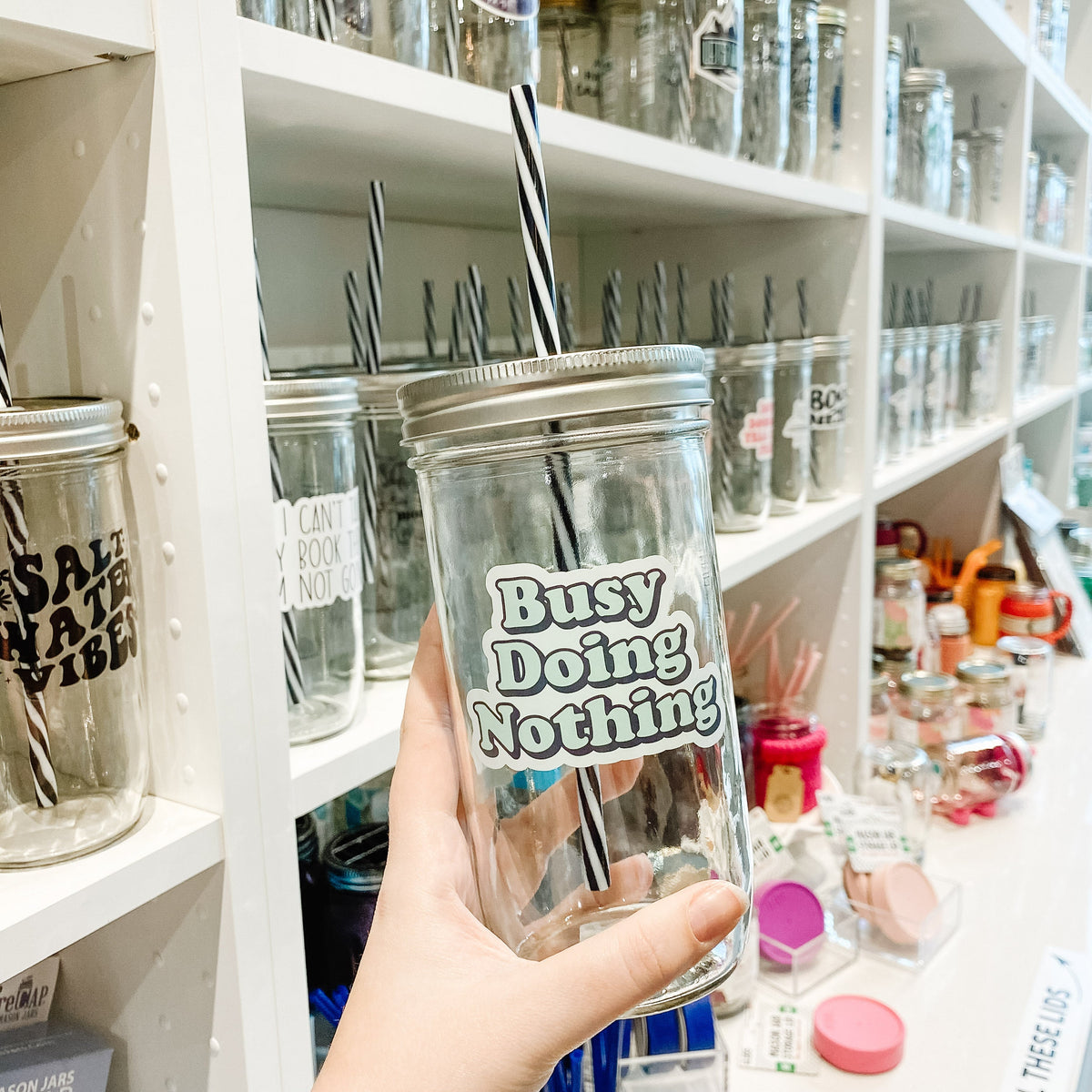 Hand holding a mason jar tumbler that says “busy doing nothing” in green letters with a black drop shadow on a white background.