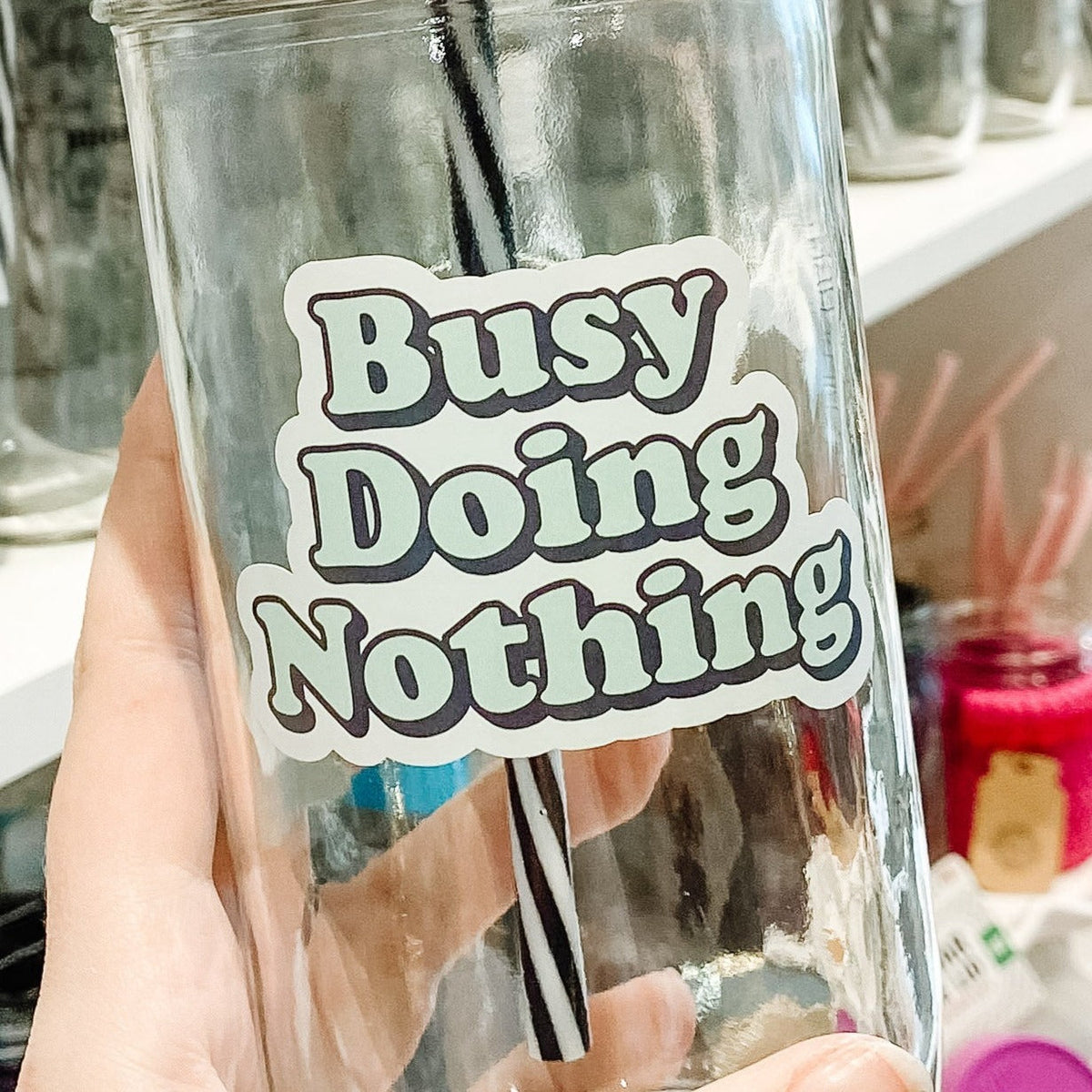 Close up of a hand holding a mason jar tumbler that says “busy doing nothing” in green letters with a black drop shadow on a white background.