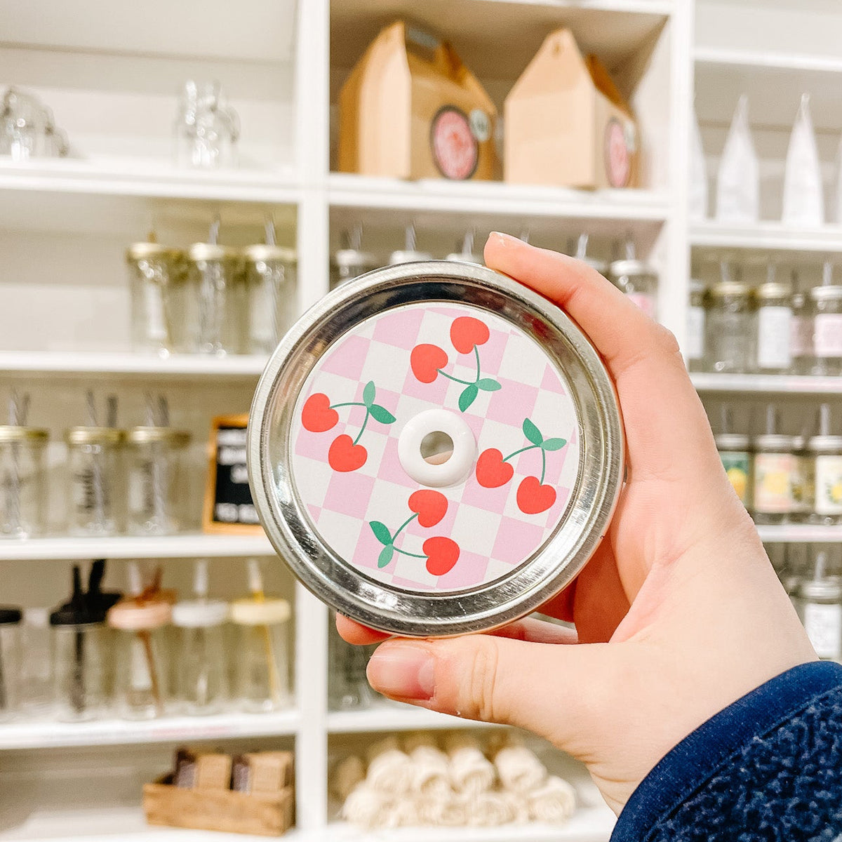 Hand holding a mason jar patterned straw lid with cherries on a white and light, pink checkered background
