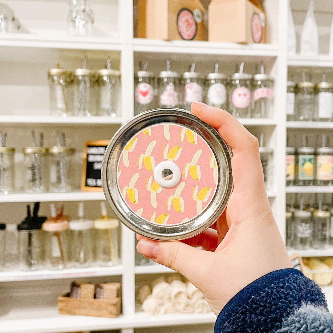 A hand holding a mason jar patterned straw lid with bananas on a pink background