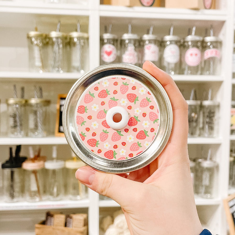 A hand holding a mason jar patterned straw lid with assorted strawberries and flowers on a light pink background
