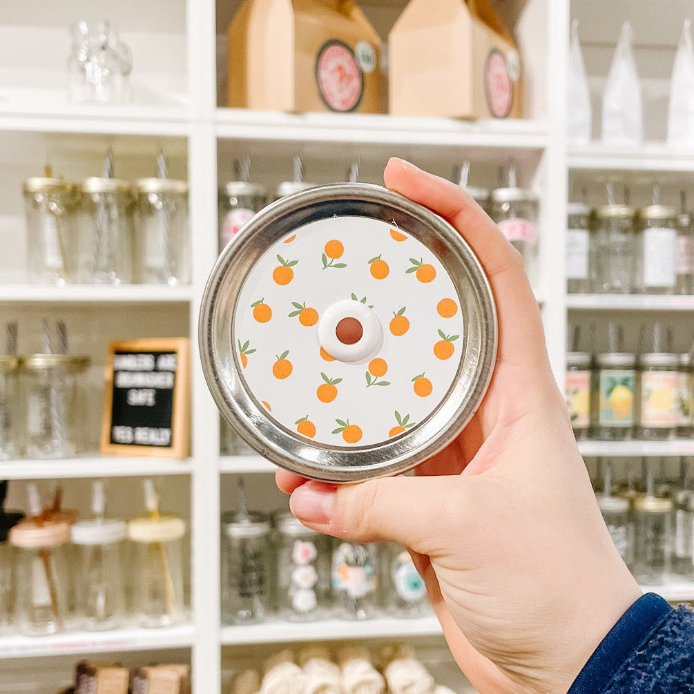 Hand holding a mason jar patterned straw lid with little oranges on a white background.