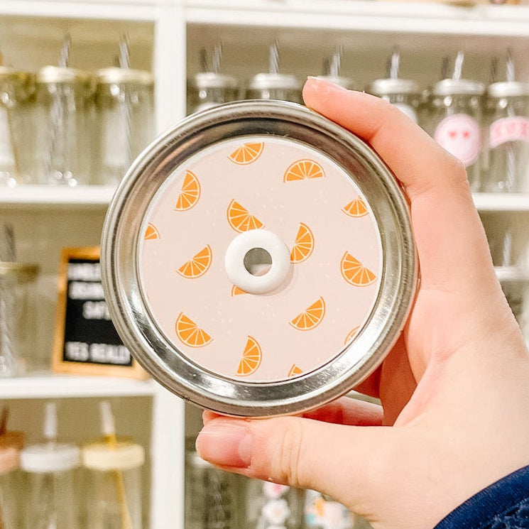 Hand holding a mason jar patterned straw lid with little orange slices on a light peach background