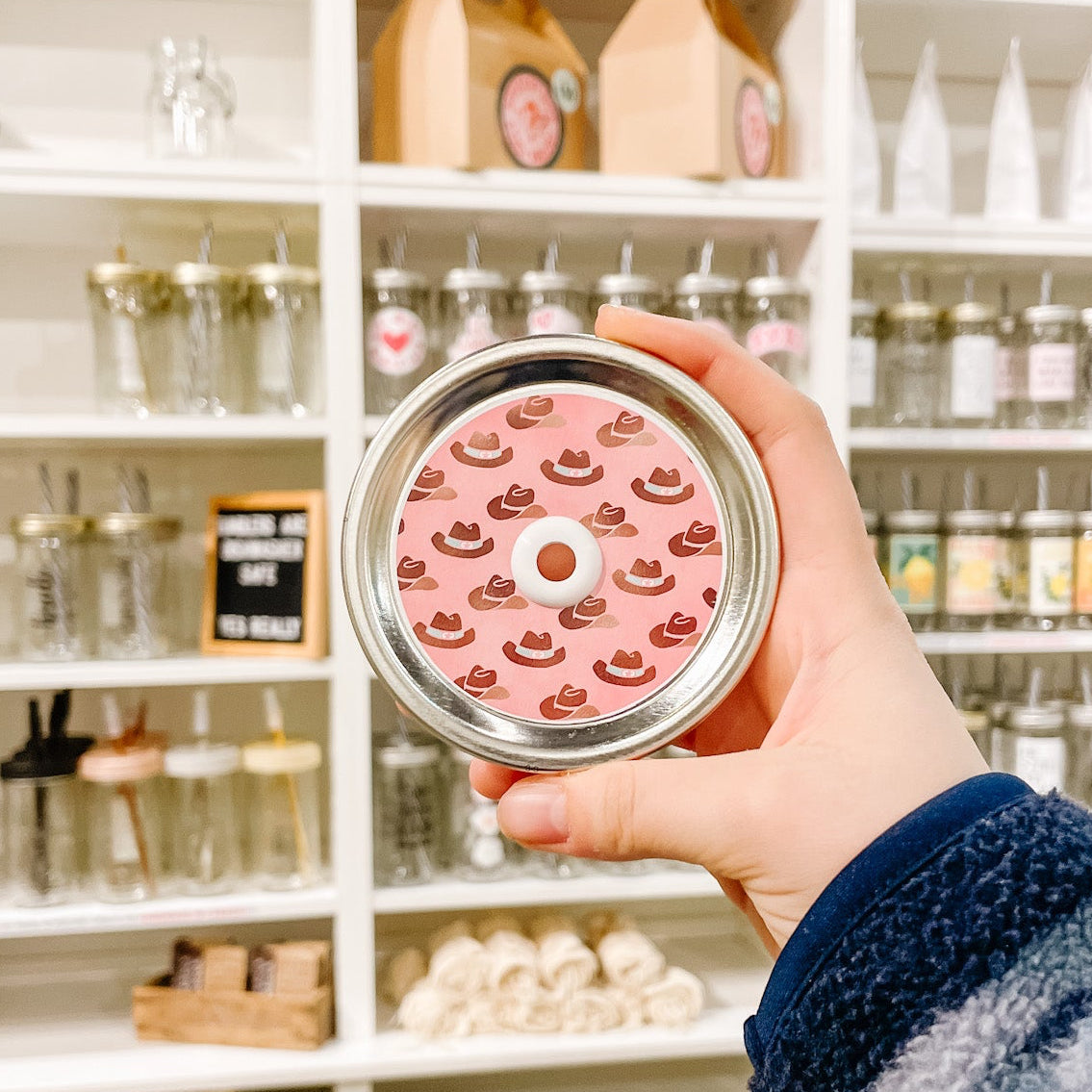 Hands holding mason jar patterned straw lid that has brown cowboy hats ona pink background