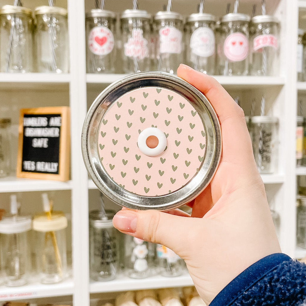 Hand holding a mason jar patterned straw lid that has small green hearts on a light pink background