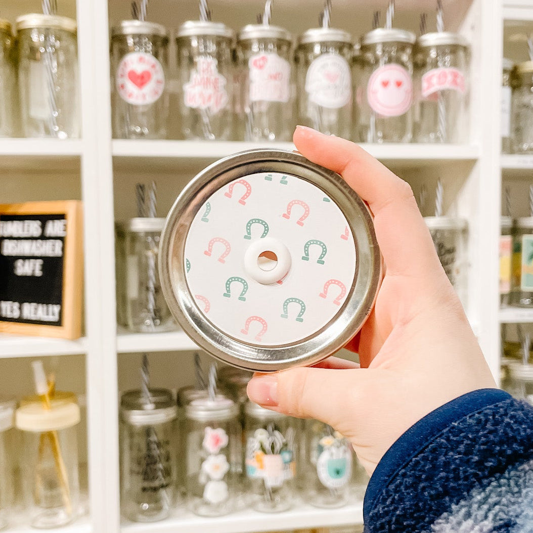 Hand holding mason jar patterned straw lid that has green and pink horseshoes on a white background.