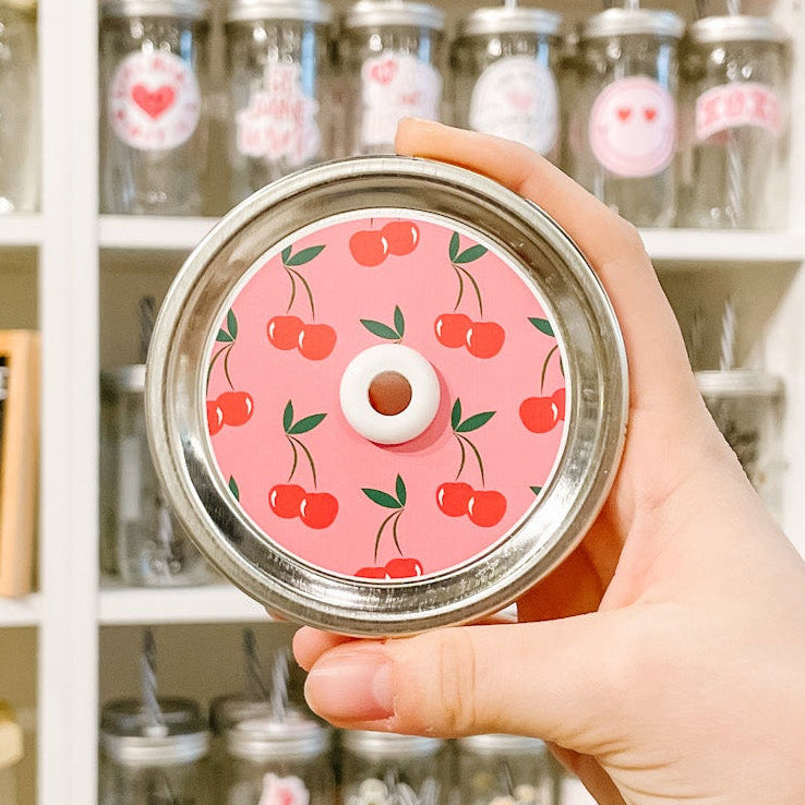 Hand holding a pink lid with cherry design in front of shelves with jars.
