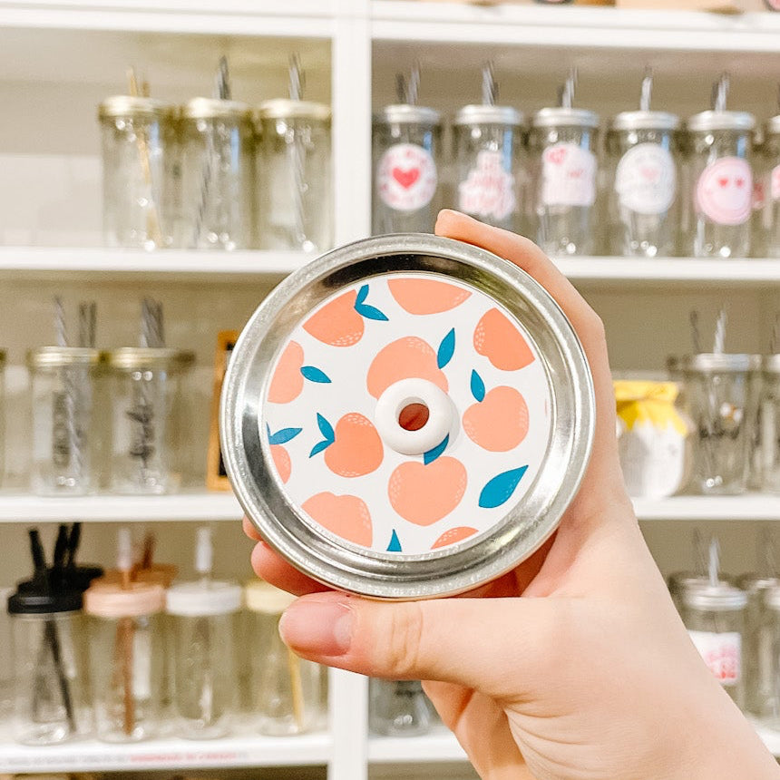 Hand holding a mason jar patterned straw lid That has peaches with teal leaves on a white background