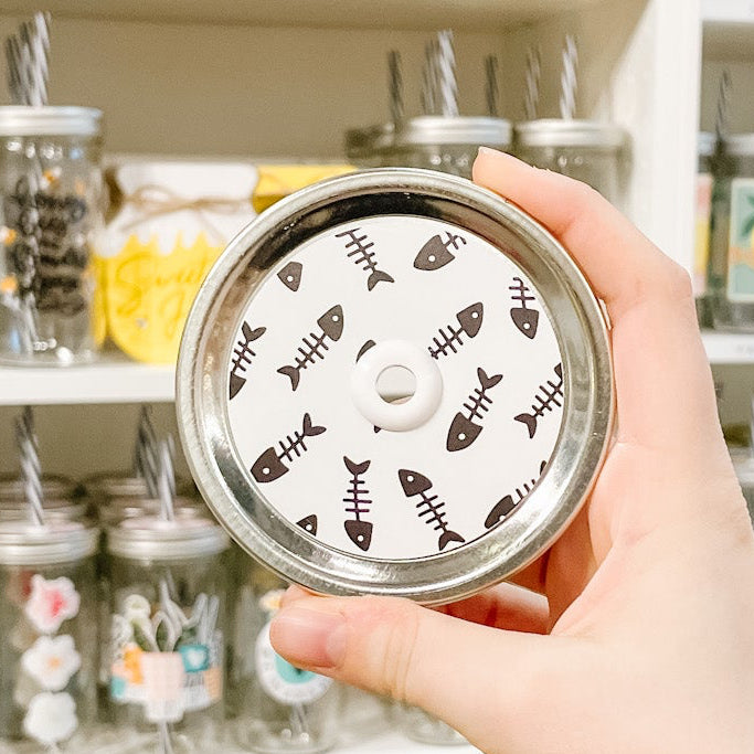 Hand holding mason jar patterned straw lid that has fish bones on white background