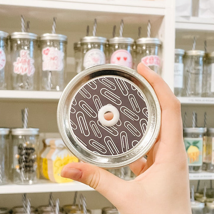 Hand holding mason jar patterned straw lid that has white paper clips on black background
