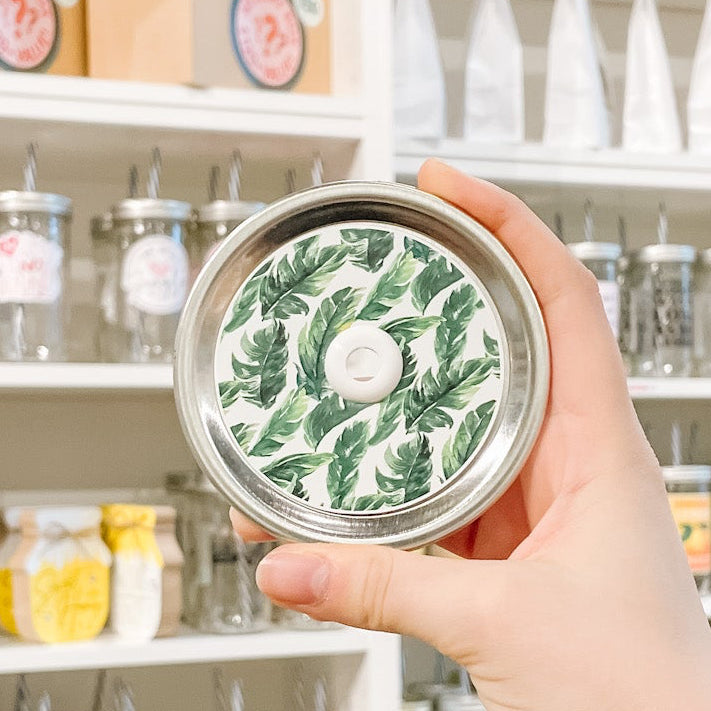 Hand holding mason jar patterned straw lid that has green tropical leaves on white background