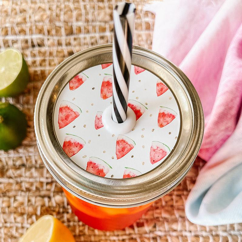 Mason jar straw lid with silver ring and a white sticker that has watermelon slices in print. Photographed as a flat lay in a weave mat with a scarf and some lemon slices.