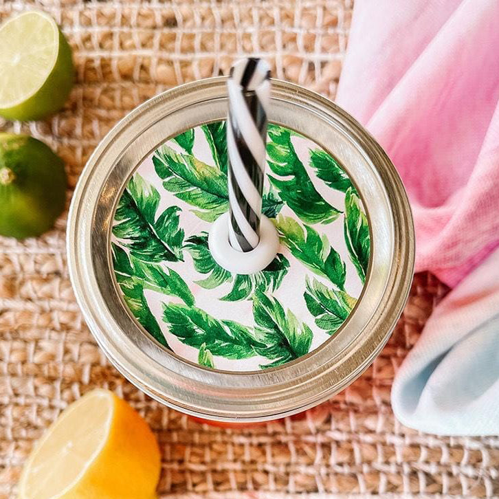 Mason jar straw lid with silver ring and a white sticker that has palm leaves in print. Photographed as a flat lay in a weave mat with a scarf and some lemon slices.