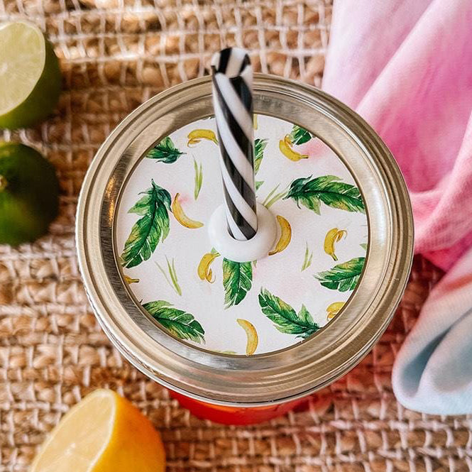 Mason jar straw lid with silver ring and a white sticker that has bananas and palm leaves in print. Photographed as a flat lay in a weave mat with a scarf and some lemon slices.