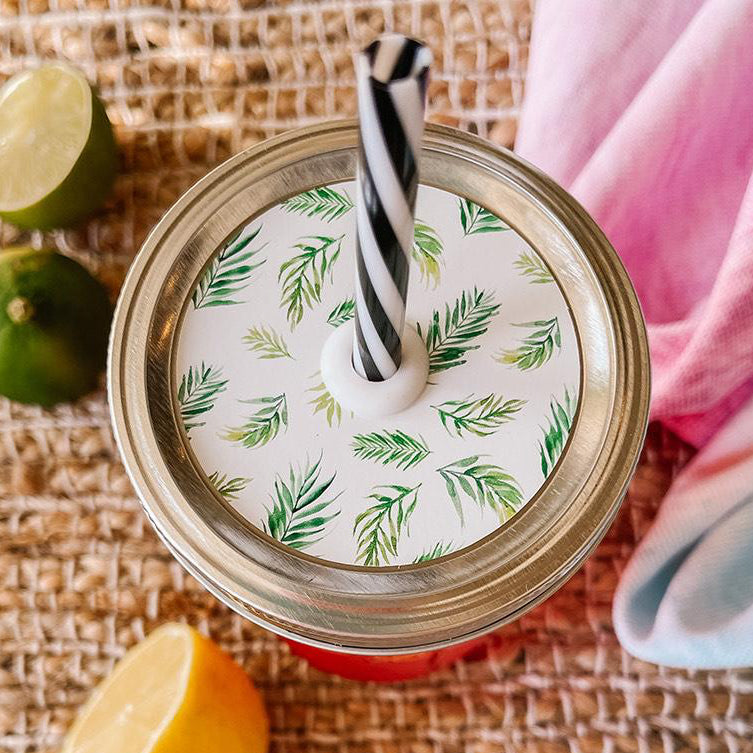 Mason jar straw lid with silver ring and a white sticker that has palm leaves in print. Photographed as a flat lay in a weave mat with a scarf and some lemon slices.