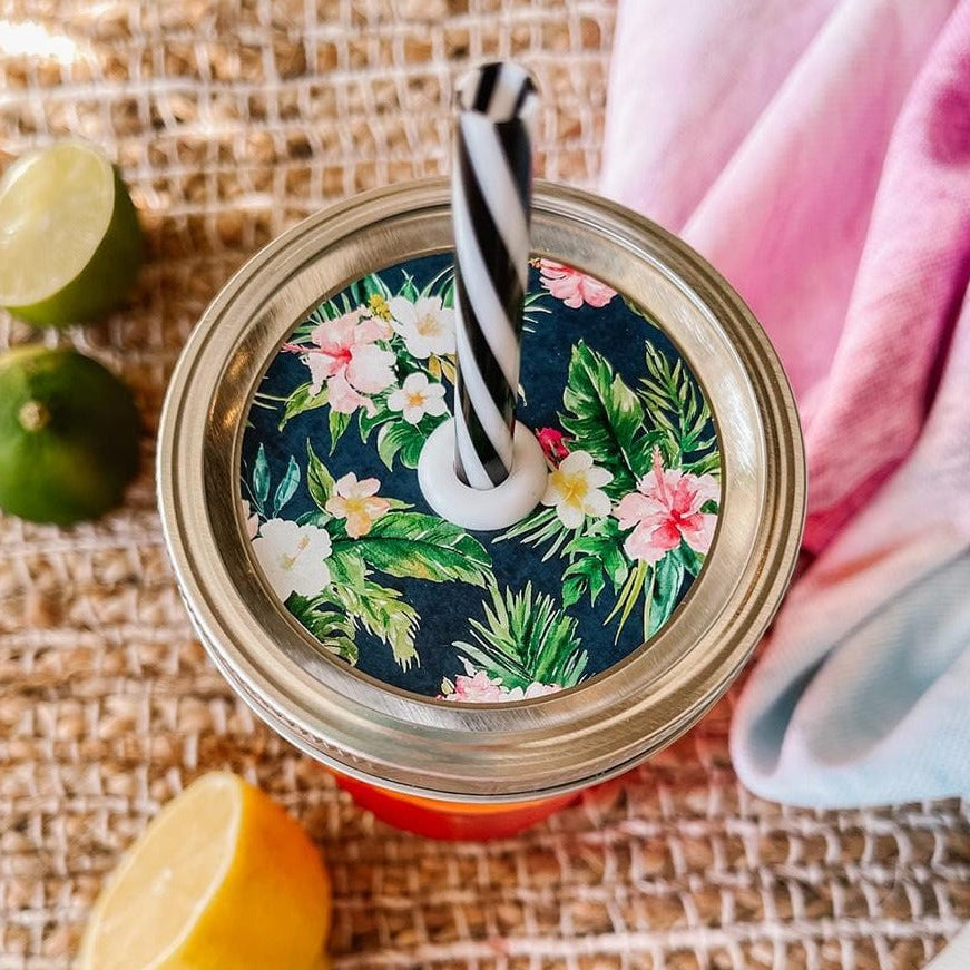 Mason jar straw lid with silver ring and a navy blue sticker that has hibiscus flowers and palm leaves in print. Photographed as a flat lay in a weave mat with a scarf and some lemon slices.