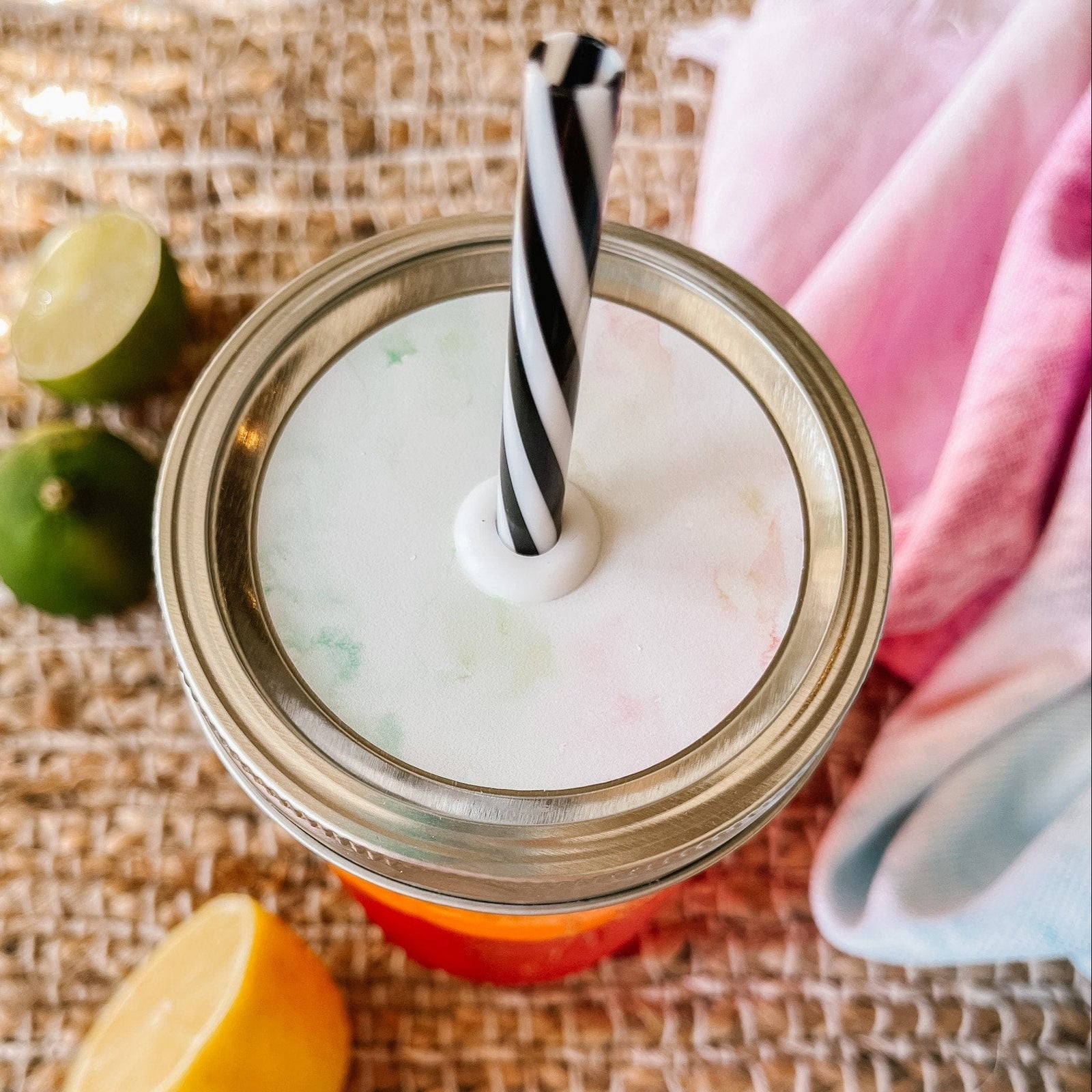 Mason jar straw lid with silver ring and a white sticker with streaks of colors of the beach and sand. Photographed as a flat lay in a weave mat with a scarf and some lemon slices.