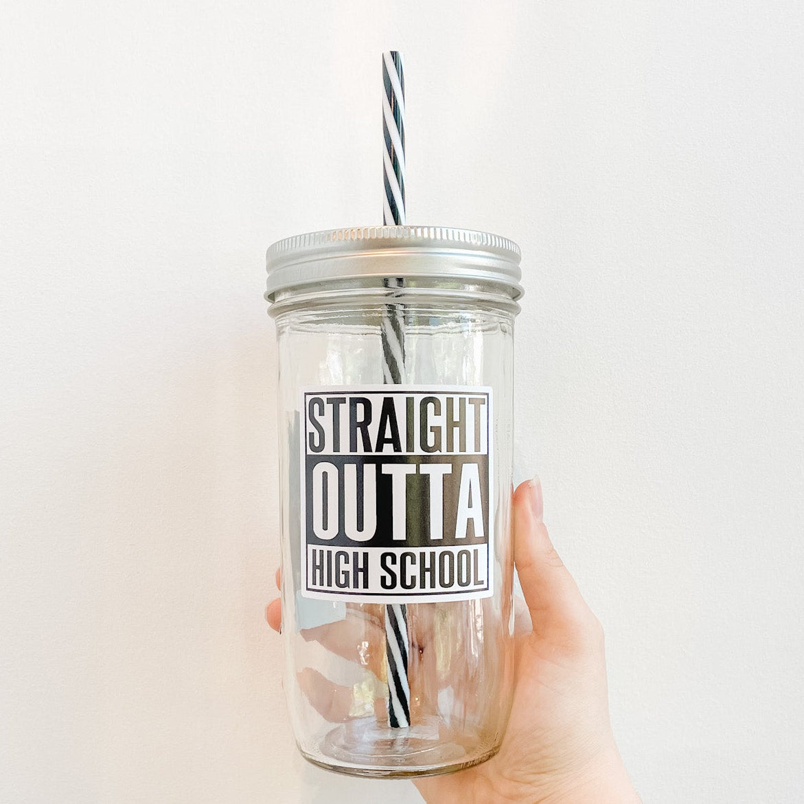 Hand holding mason jar tumbler that reads in bold black and white text straight outta high school. Reusable black and white striped straw and silver straw lid.
