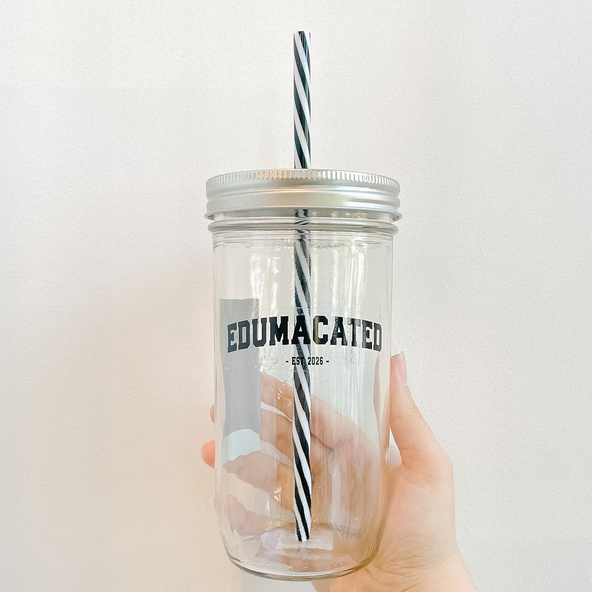 Hand holding mason jar tumbler that reads edumacated est 2026 in black text and has reusable black and white striped straw and straw lid
