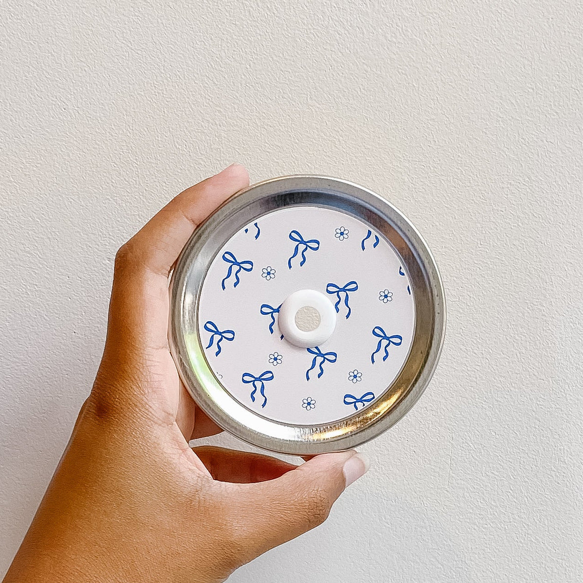 Hand holding mason jar patterned straw lid that has little blue bows and flower designs on white