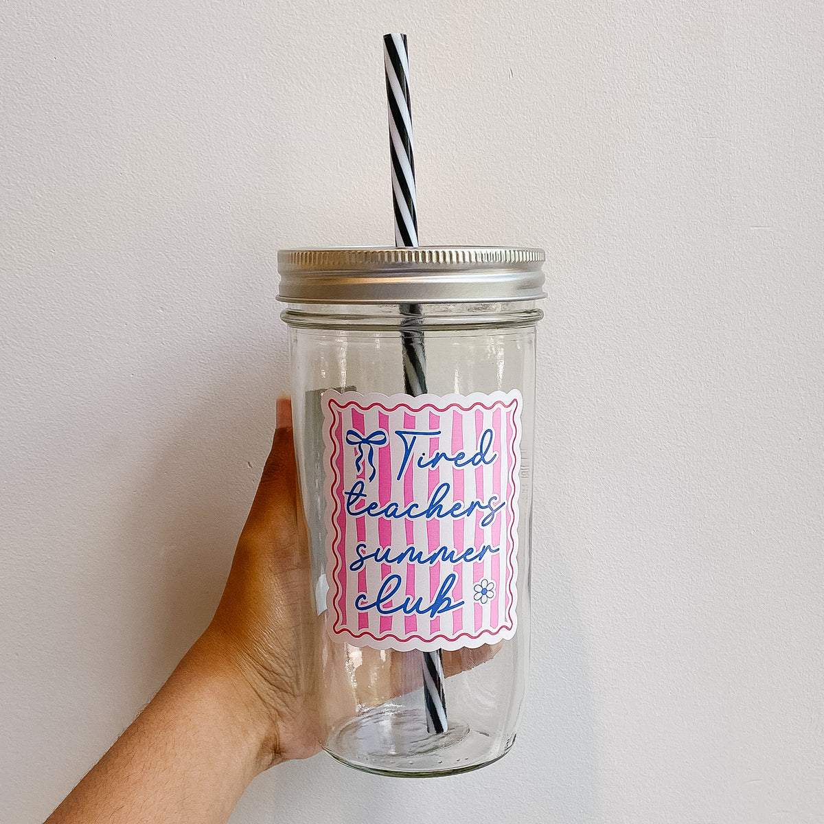 Hand holding a mason jar tumbler that reads tired teachers summer club in blue ink with white red and pink striped design on background