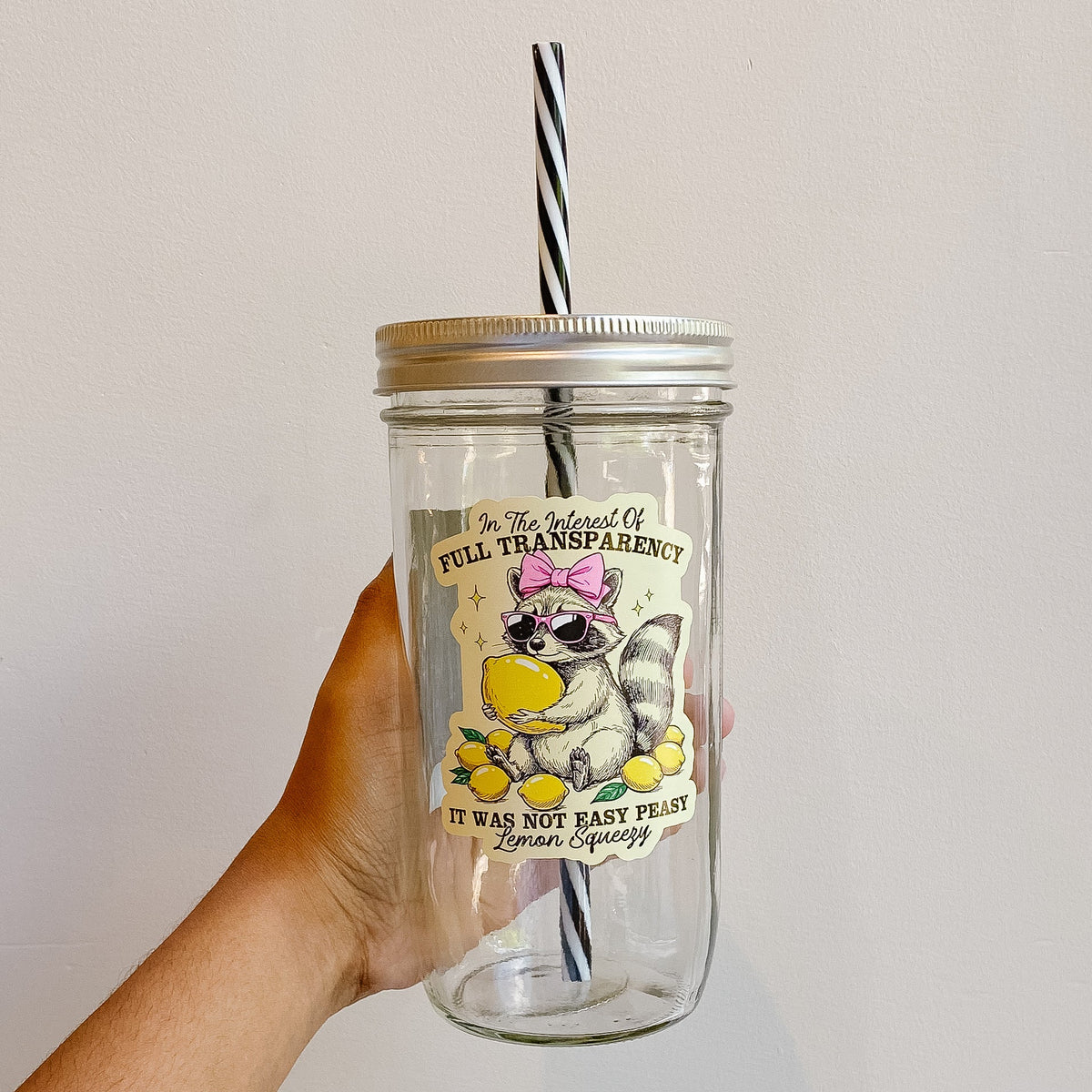 Hand holding a mason jar tumbler with a funny raccoon design holding lemons and wearing a pink bow and sunglasses surrounded by lemons. Black and white striped straw.