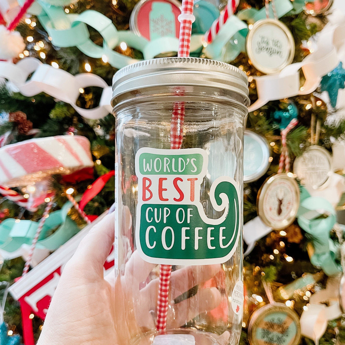 A woman holding an Elf Theme regular mouth mason jar tumbler with silver lid and red and white pattern straw. The tumbler has a print that says "World's Best Cup of Coffee" in a red and green color. Behind it is a Christmas tree.