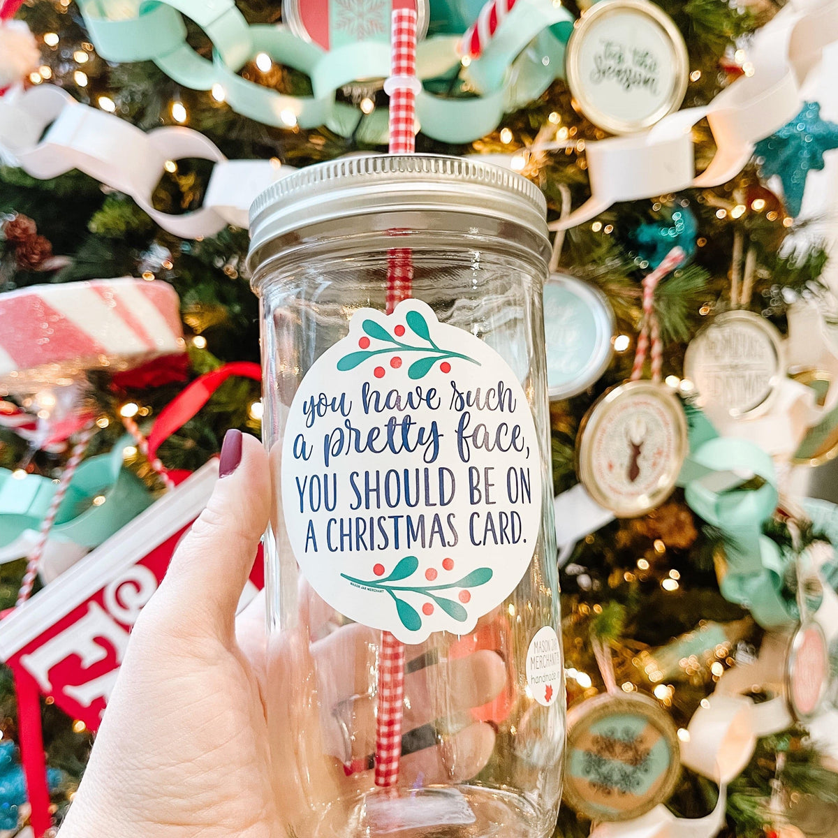 A woman holding an Elf Theme regular mouth mason jar tumbler with silver lid and red and white pattern straw. The tumbler has a print that says "You have such a pretty face, You should be on a Christmas card" in green. Behind it is a Christmas tree.