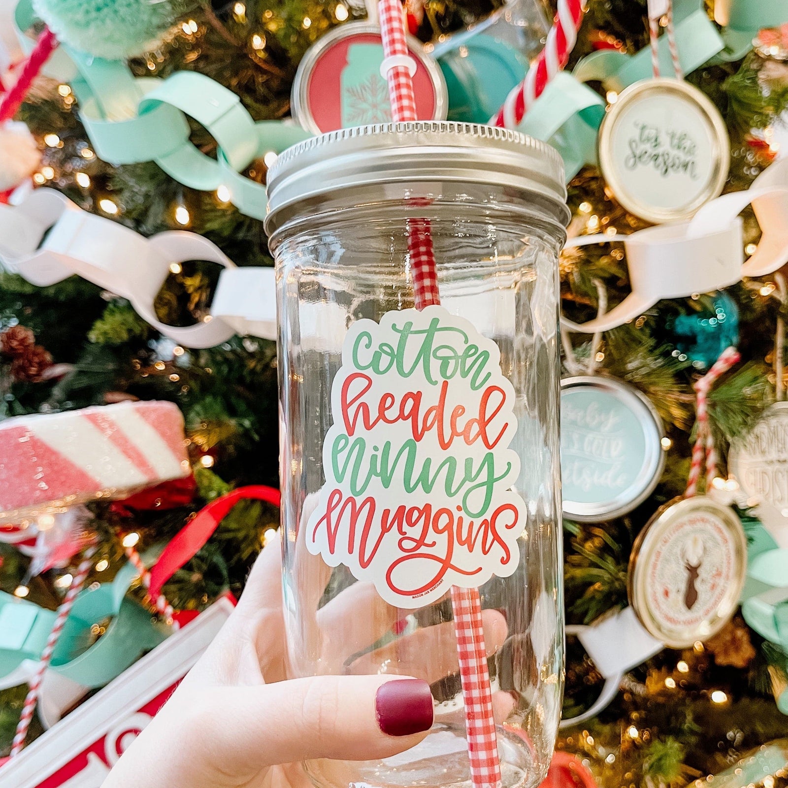A woman holding an Elf Theme regular mouth mason jar tumbler with silver lid and red and white pattern straw. The tumbler has a print that says "cotton headed Ninny Muggins" in a red and green color. Behind it is a Christmas tree.