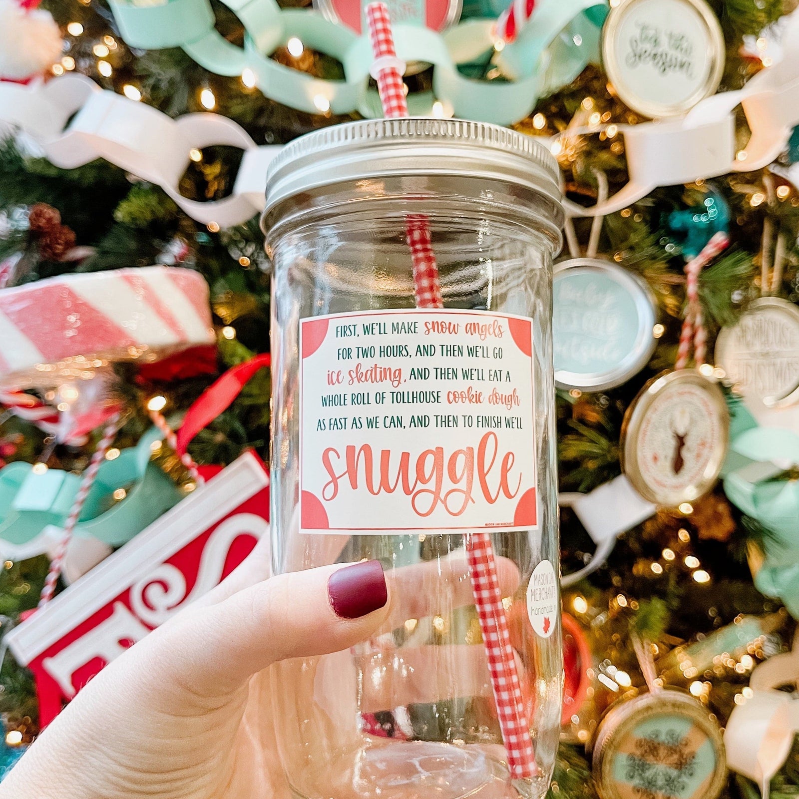 A woman holding an Elf Theme regular mouth mason jar tumbler with silver lid and red and white stripe straw. The tumbler has a print that says "Snuggle" and behind it is a Christmas tree. The tumbler has a red and white patterned color.