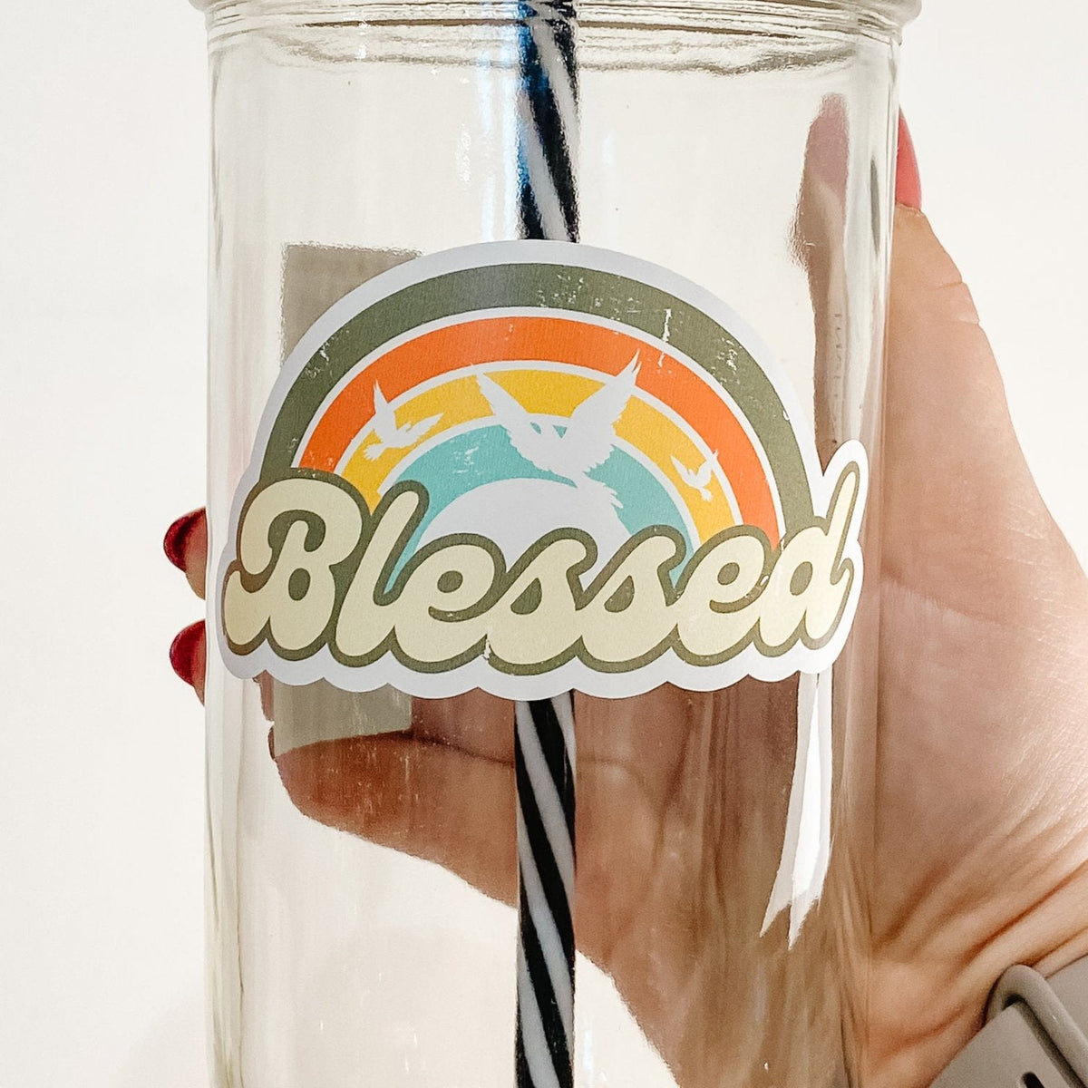 Close up of a hand holding a mason jar tumbler that says "Blessed" with a rainbow and white silhouettes of birds.