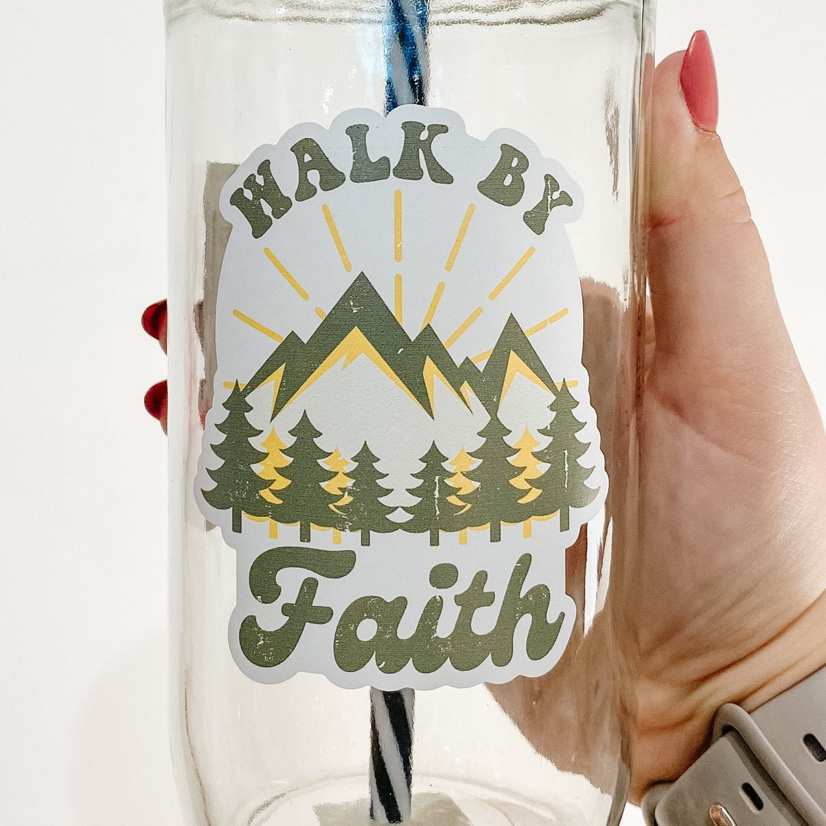 Close up of a hand holding a mason jar tumbler that says "walk by faith" with mountains and trees (green and yellow).