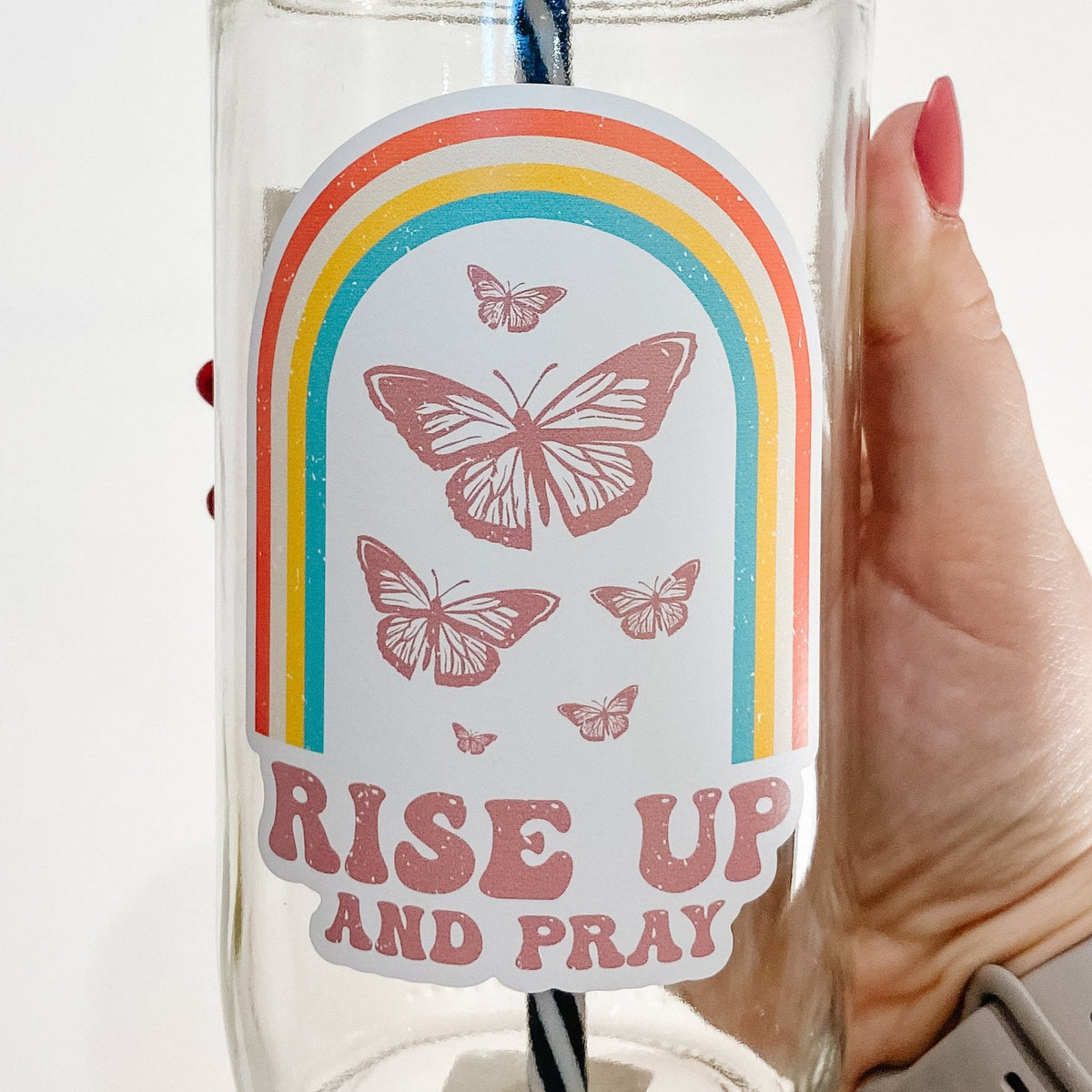Close up of hand holding a mason jar tumbler that says "Rise up and pray" with a rainbow and butterflies above the words