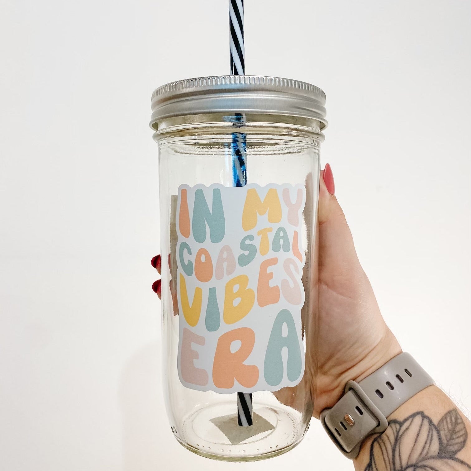 hand holding a mason jar tumbler that reads "in my coastal vibes era" the lettering is bubbly and in different shades of yellow, orange, blue, and pink