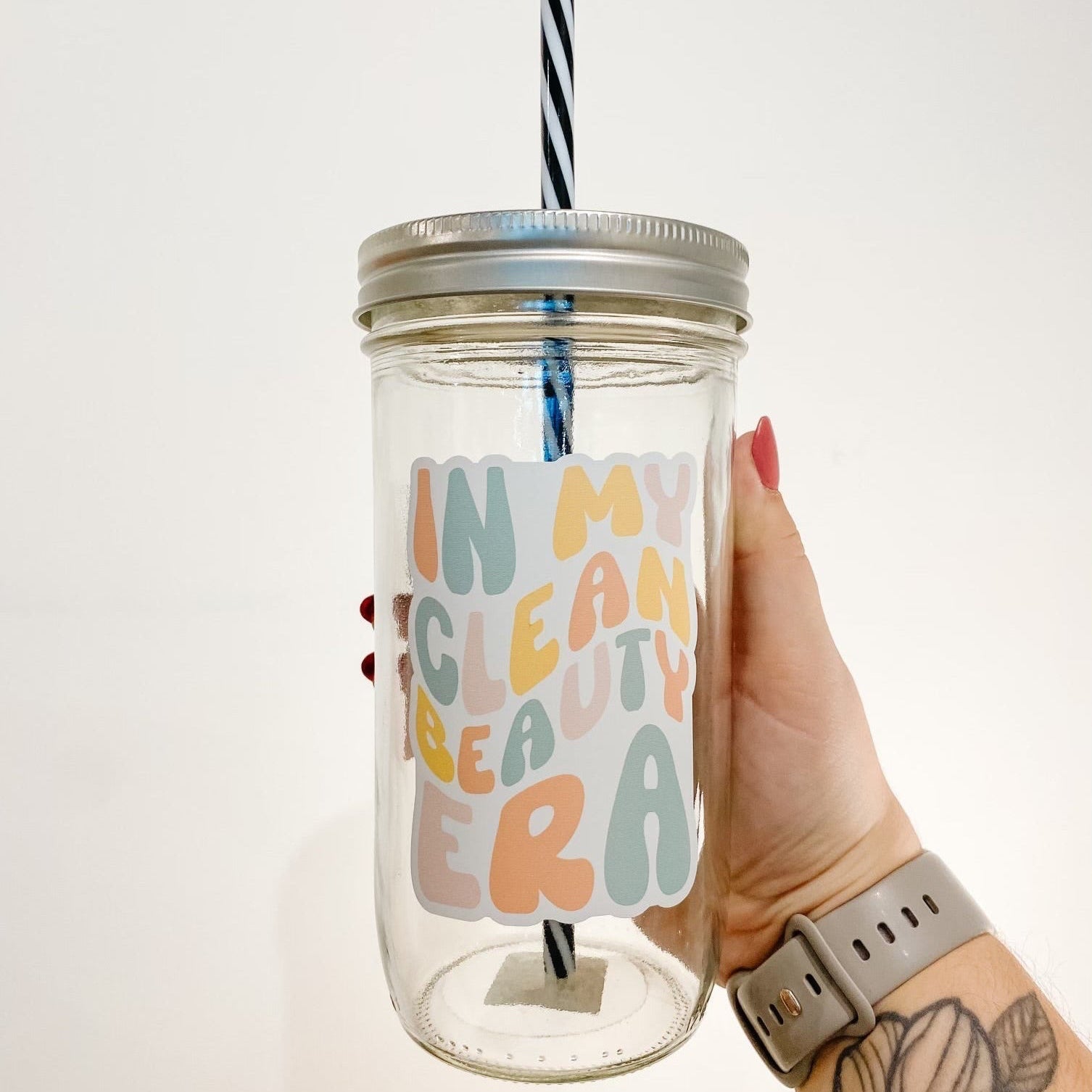 hand holding a mason jar tumbler that has a decal that reads "in my clean beauty era" the lettering is bubbly and in different shades of orange, yellow, pink, and blue