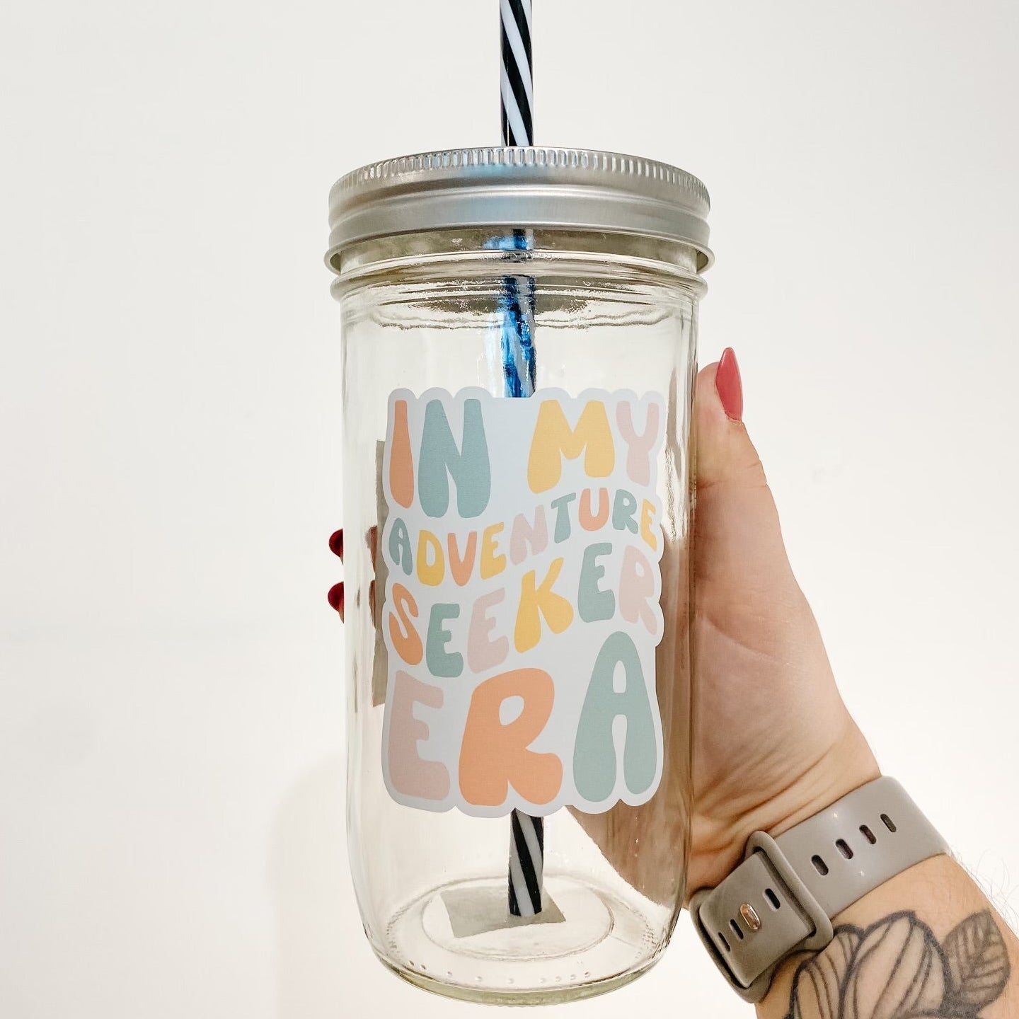 hand holding a mason jar tumbler that reads "in my adventure seeker era" the lettering is bubbly and in different shades of orange, yellow, pink, and blue