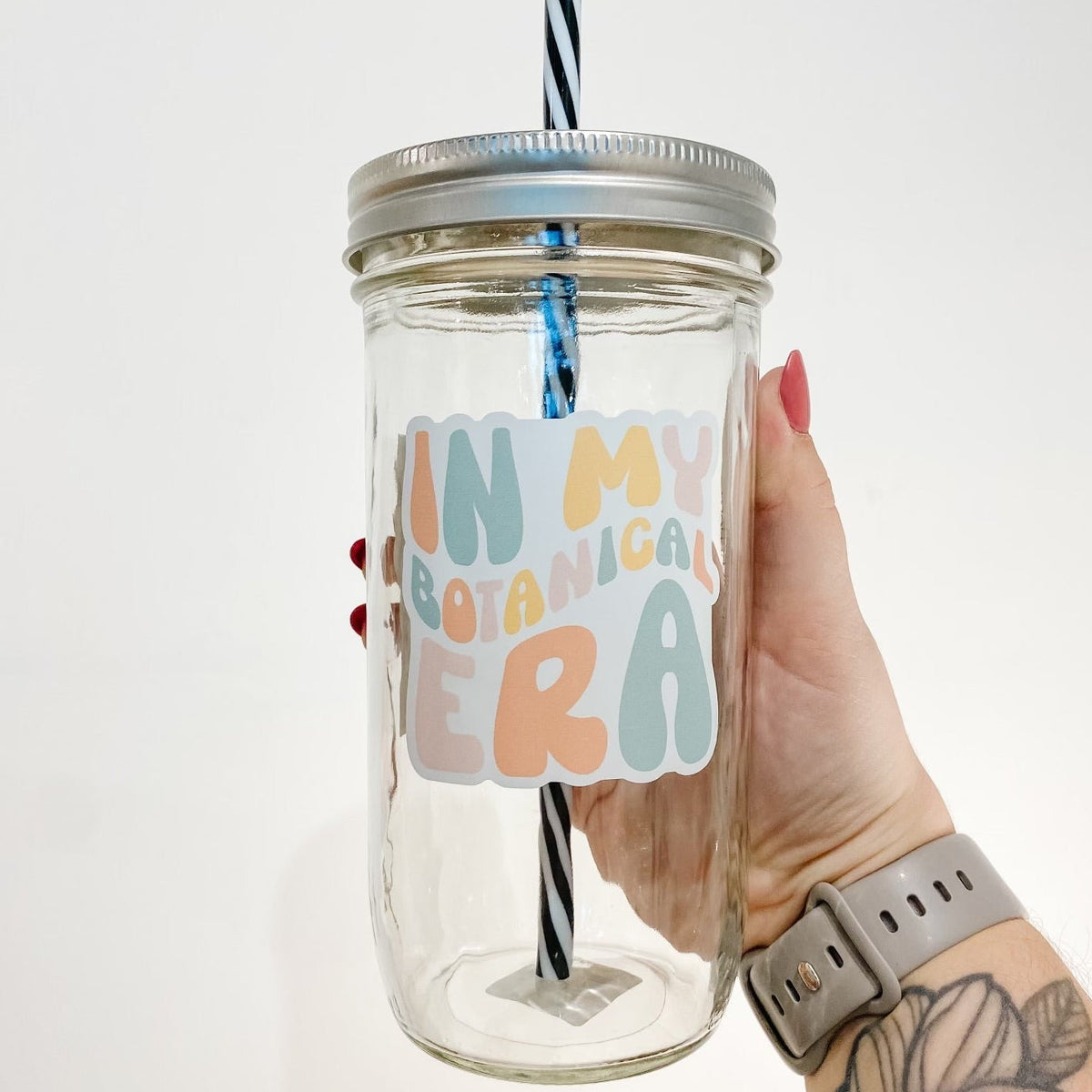 hand holding a mason jar tumbler that says "in my botanical era" the lettering is bubbly and in different shades of orange, yellow, pink, and blue