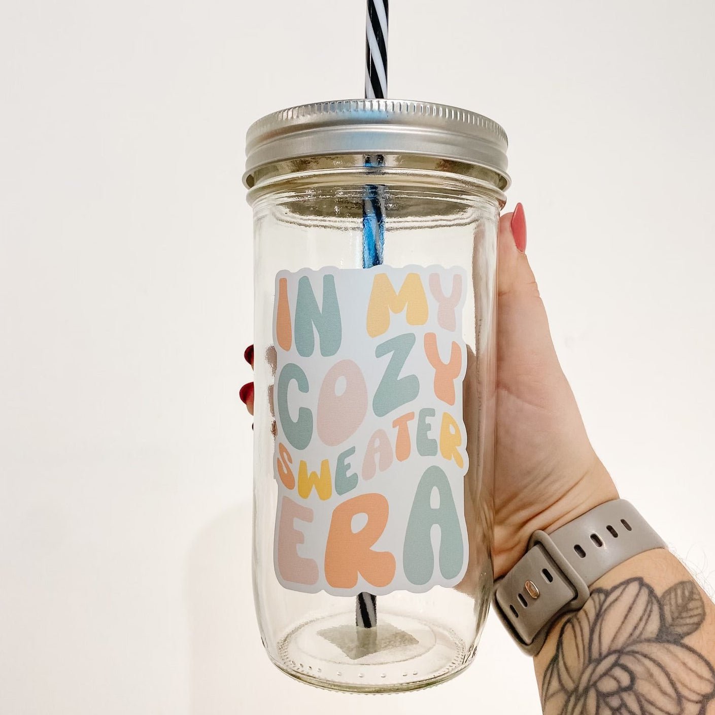 hand holding a mason jar tumbler that reads "in my cozy sweater era" the lettering is bubbly and in different shades of orange, yellow, pink, and blue