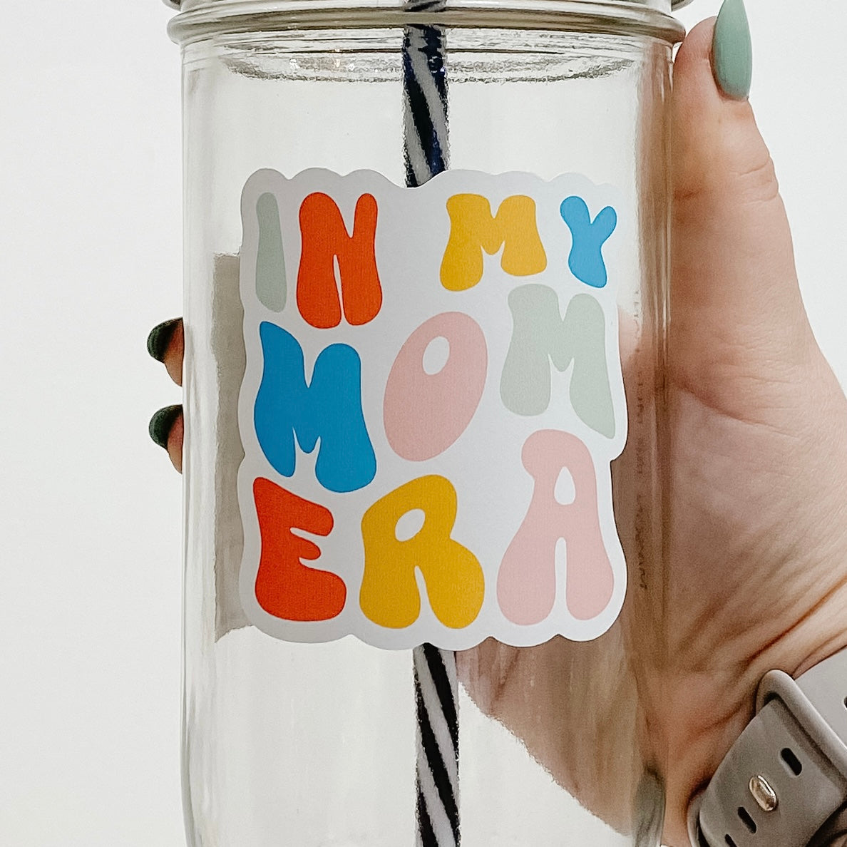 Close up of a hand holding a msaon jar tumbler that says "In my mom era" in bubbly, different colored letters.