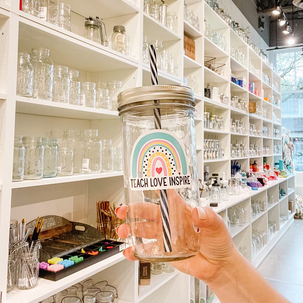 Photo of a mason jar tumbler with gold lid and black and white straw. Tumbler has a sticker with a rainbow design and a heart in the middle. Underneath is a print that reads, "Teach Love Inspire."