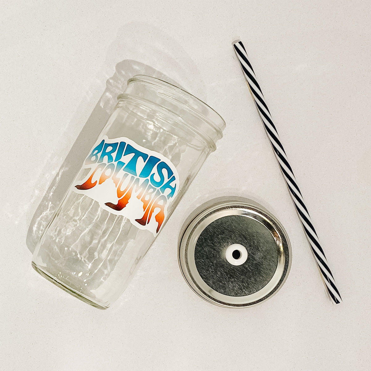 Flat lay of mason jar tumbler that reads British Columbia in shape of bear. Colours are muted sunset gradient. Black and white striped straw. Mason jar straw lid is plain silver.