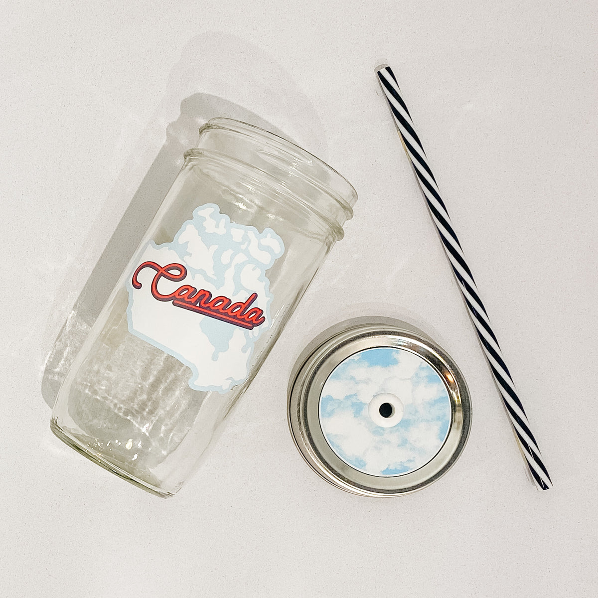 Flat lay of mason jar tumbler that reads Canada in red surrounded by blue shape of Canada. Black and white striped straw. Mason jar straw lid is clouds with blue background.