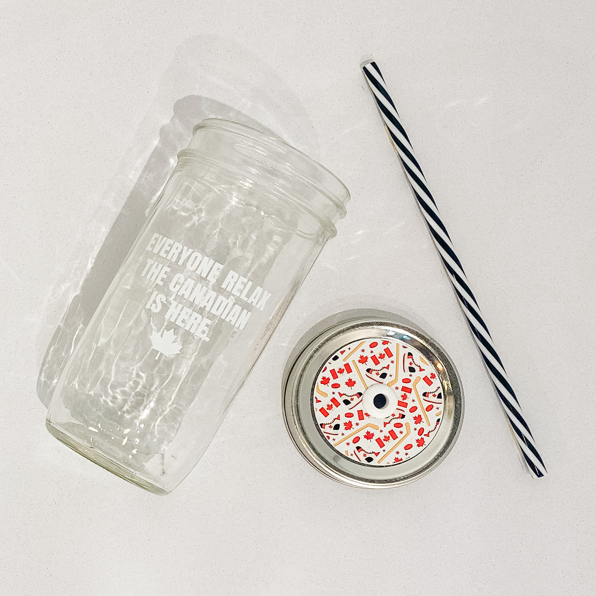 Flat lay off Mason jar tumbler that reads everyone relax the Canadian is here in white text with small maple leaf at bottom. Black and white striped straw. Mason jar straw lid is Canada flag, hockey sticks, ice skates and maple leafs on white background.